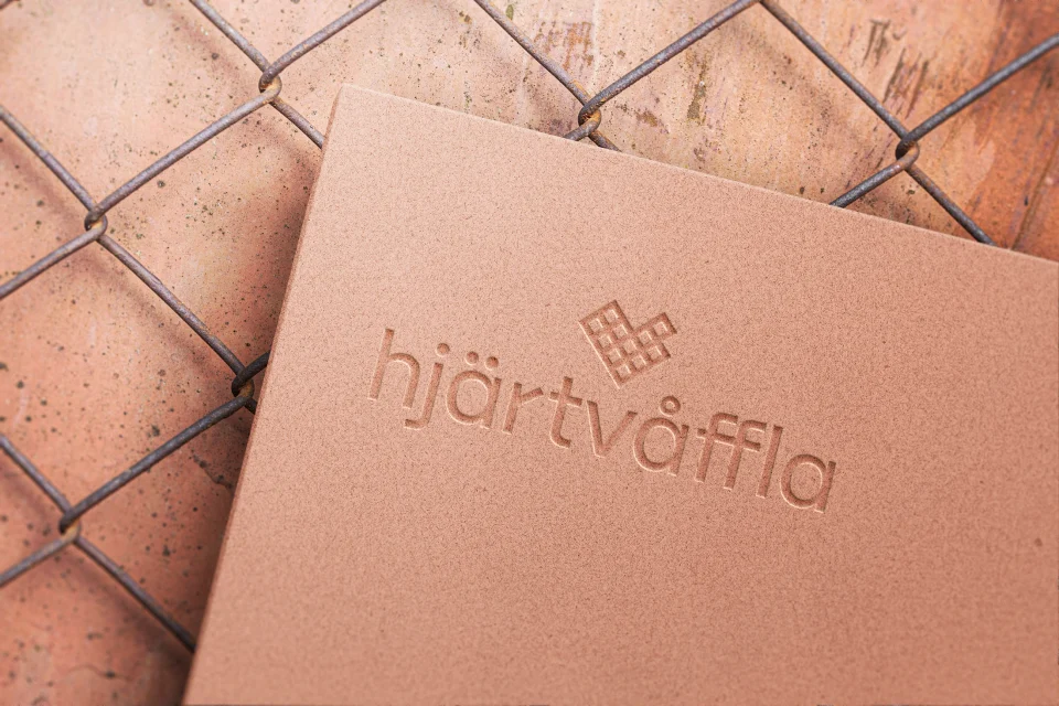 The hjärtvåffla wordmark and heart-shaped waffle symbol debossed into peach-coloured card on a terracotta surface.