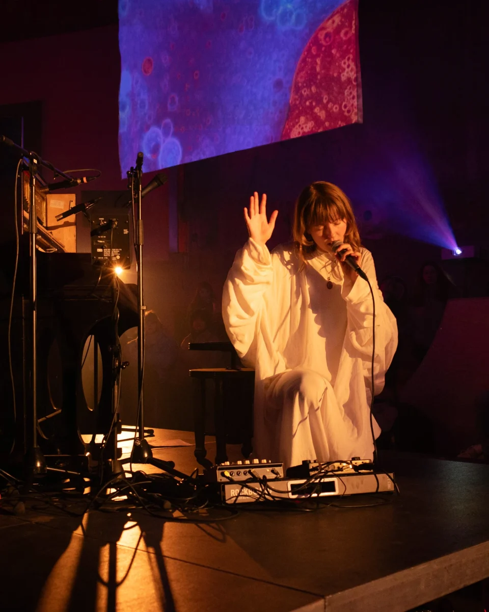A seated singer performs beneath microscopic projections, surrounded by instruments and stage equipment.