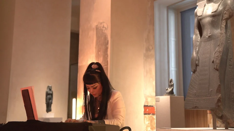 Lyra von Rose plays a keyboard among Egyptian sculptures and warmly lit columns at Neues Museum.