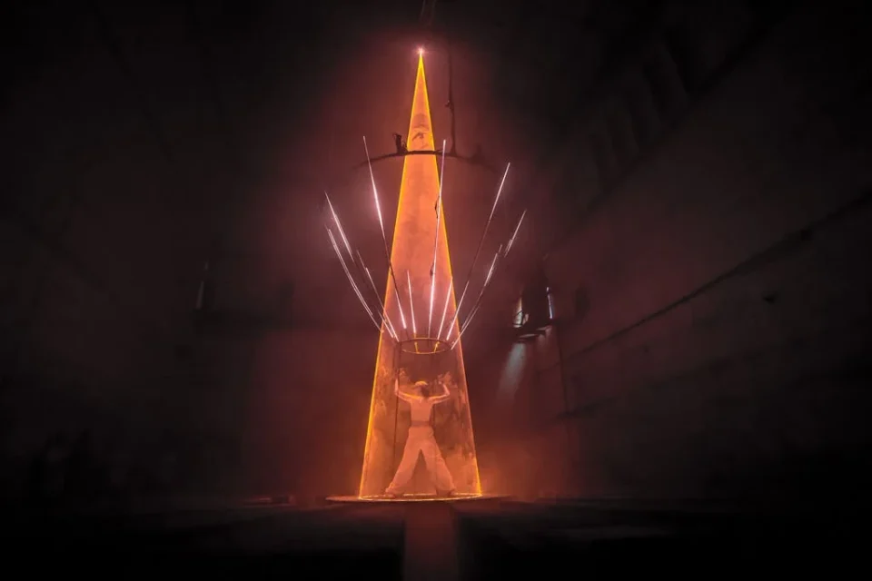A visitor stands beneath a narrow orange cone of laser light in the industrial hall at Monopol.
