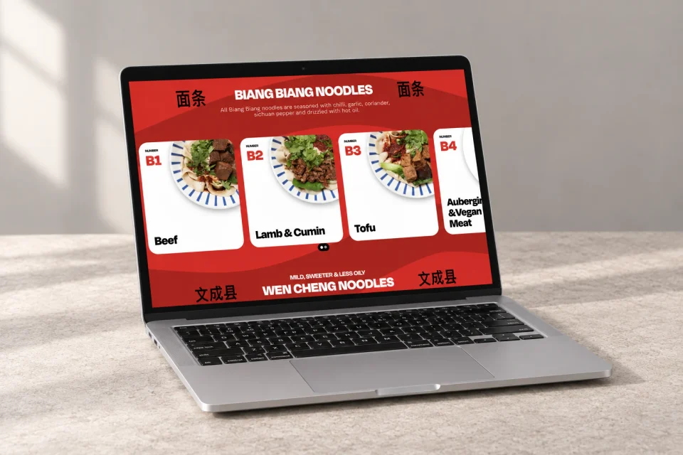 A silver laptop on pale stone showing the red Biang Biang noodle menu and illustrated dish cards.
