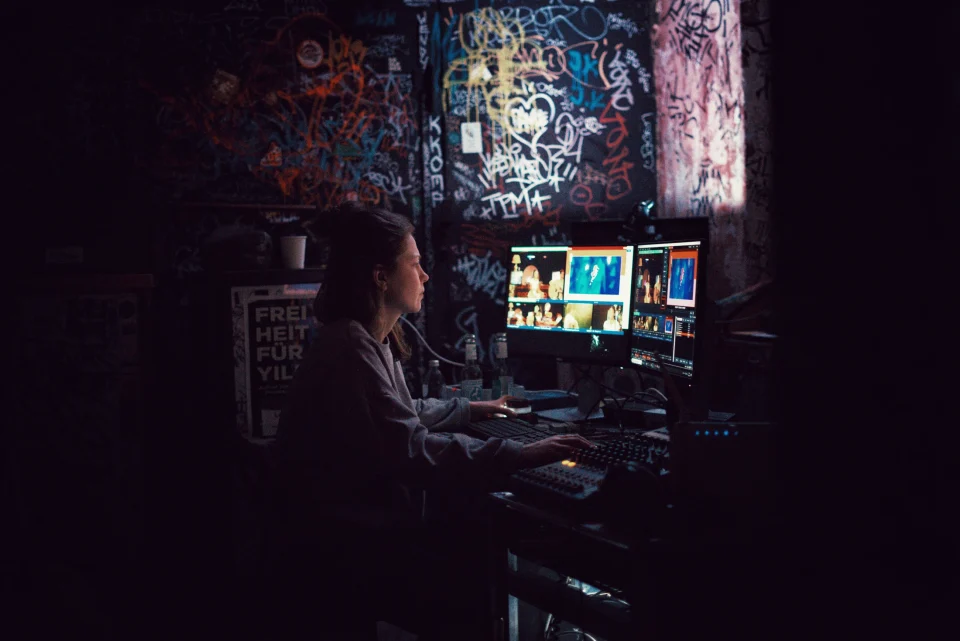 A person working at a multi-monitor broadcast station inside a dark, poster-lined club.