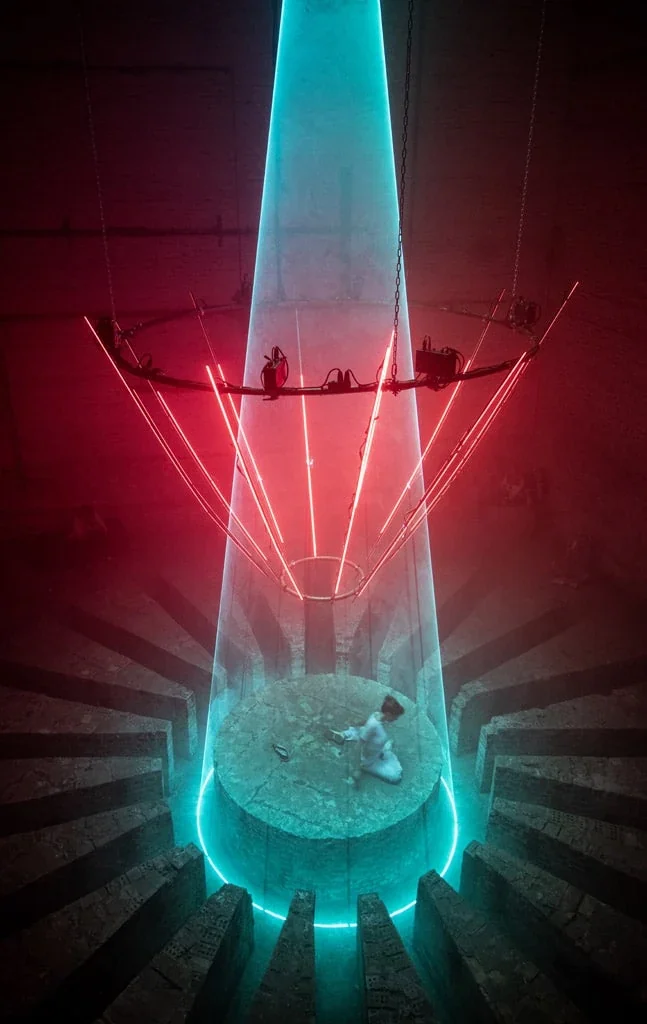 An elevated view of turquoise laser light, red illuminated structures and a visitor beneath the INVERSION installation.